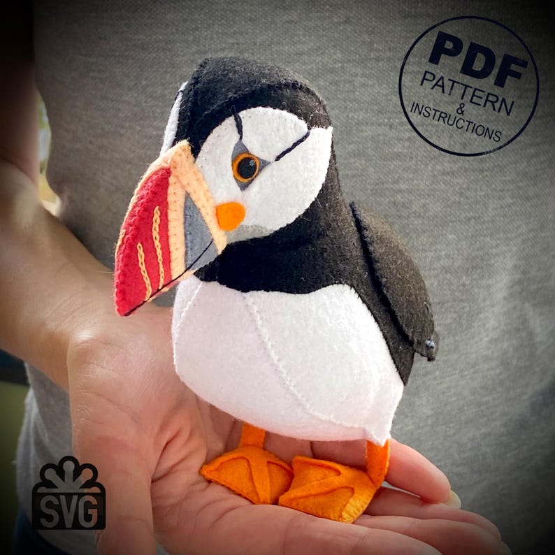 Peut inclure: Un jouet en peluche en forme de macareux, en feutre, avec un bec rouge, orange et blanc. Le macareux est assis dans la main d'une personne. L'image comprend le texte "PDF PATTERN & INSTRUCTIONS" et "SVG".