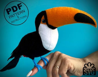 May include: A felt toucan bird with a black body, white throat, and orange beak. The toucan is perched on a finger with a blue ribbon tied around it. The image includes the text "PDF PATTERN & INSTRUCTIONS" and "SVG".