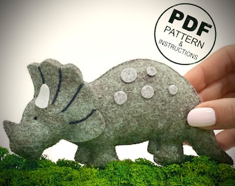 May include: A gray felt triceratops dinosaur toy with white felt spots. The toy is sitting on a bed of green moss. The image includes the text "PDF PATTERN & INSTRUCTIONS".