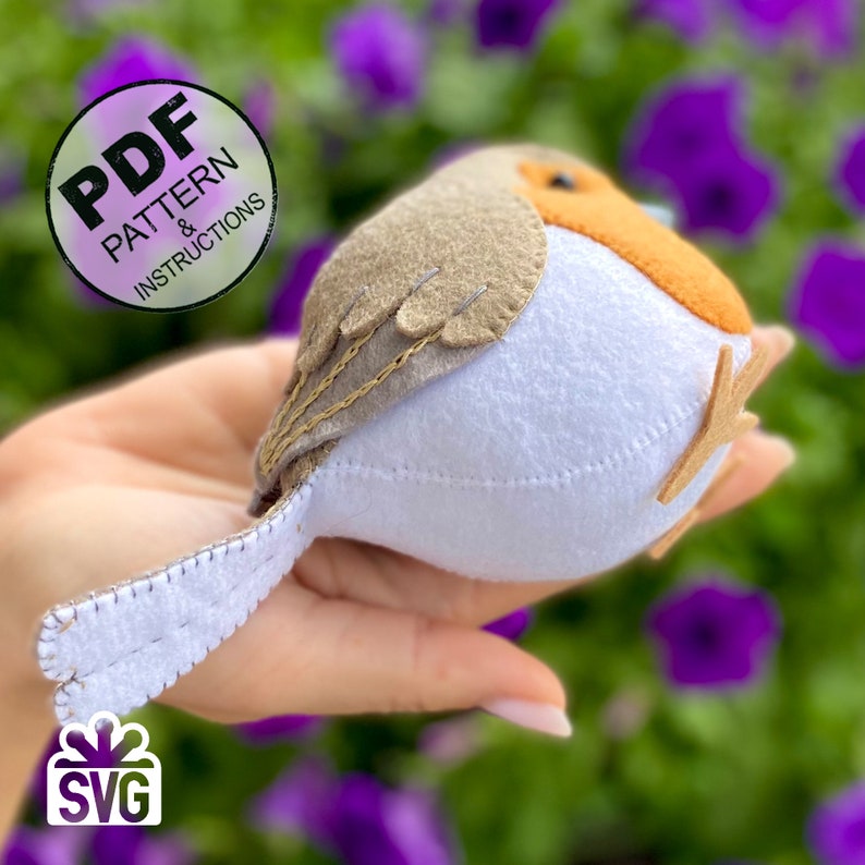 K&ouml;nnte beinhalten: Ein Filz-Rotkehlchen mit wei&szlig;em Bauch, braunem R&uuml;cken und orangefarbener Brust. Der Vogel ist mit braunem Garn gen&auml;ht und hat einen kleinen braunen Filzschwanz. Der Vogel wird in einer Hand vor einem Hintergrund aus lila Blumen gehalten.