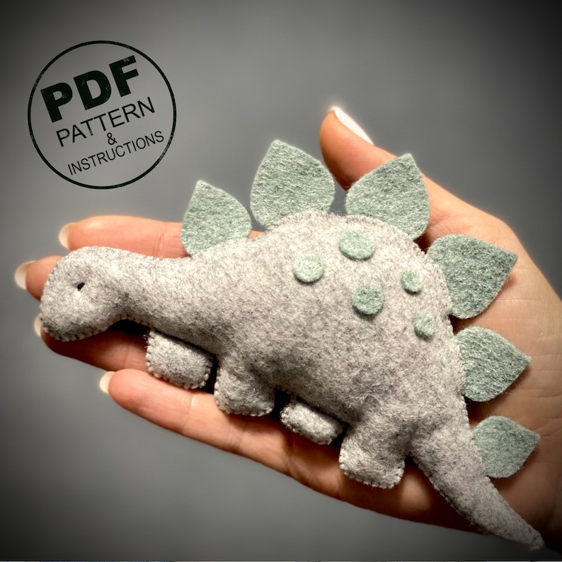 K&ouml;nnte beinhalten: Ein grauer Filzdinosaurier mit gr&uuml;nen Filzstacheln auf dem R&uuml;cken. Der Dinosaurier wird in einer Hand gehalten. Der Text "PDF PATTERN & INSTRUCTIONS" befindet sich in einem Kreis auf dem Bild.