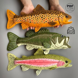 Puede incluir: Tres peces de fieltro hechos a mano con costuras detalladas. El pez superior es naranja con manchas marrones, el del medio es verde oliva con vientre blanco y el inferior es verde claro con una franja lateral rosa y manchas negras.