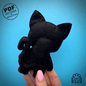 May include: A handmade black felt cat toy, viewed from the back, with a curled tail and pointed ears. The cat is on a blue background. The image includes the text "PDF PATTERN & INSTRUCTIONS" in a circle.