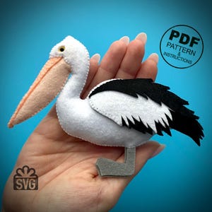 May include: A white felt pelican with a black and white wing, a pink beak, and grey feet. The pelican is being held in a hand. The image includes the text "PDF PATTERN & INSTRUCTIONS" and "SVG".