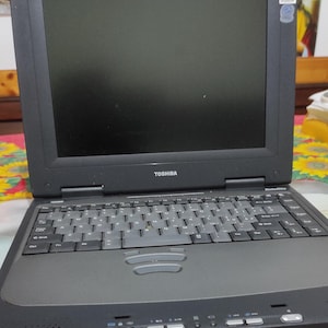 Peut inclure: Un ordinateur portable Toshiba Satellite noir avec un clavier, un écran et le logo Toshiba. L'ordinateur portable est ouvert et posé sur une surface à motifs floraux. Un autocollant Windows est visible sur l'écran.