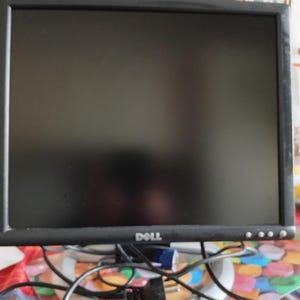 Pode incluir: Um monitor de computador Dell preto com uma base prateada. O monitor tem uma moldura preta e um ecrã escuro. O logótipo da Dell é visível na parte inferior central. Os cabos estão ligados à parte traseira do monitor.
