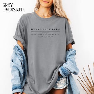 May include: A gray oversized t-shirt with the text "HURKLE-DURKLE" in black. The text is followed by "SCOTTISH" and a definition: "TO LAY AROUND IN BED LONG AFTER YOU SHOULD HAVE GOT UP".