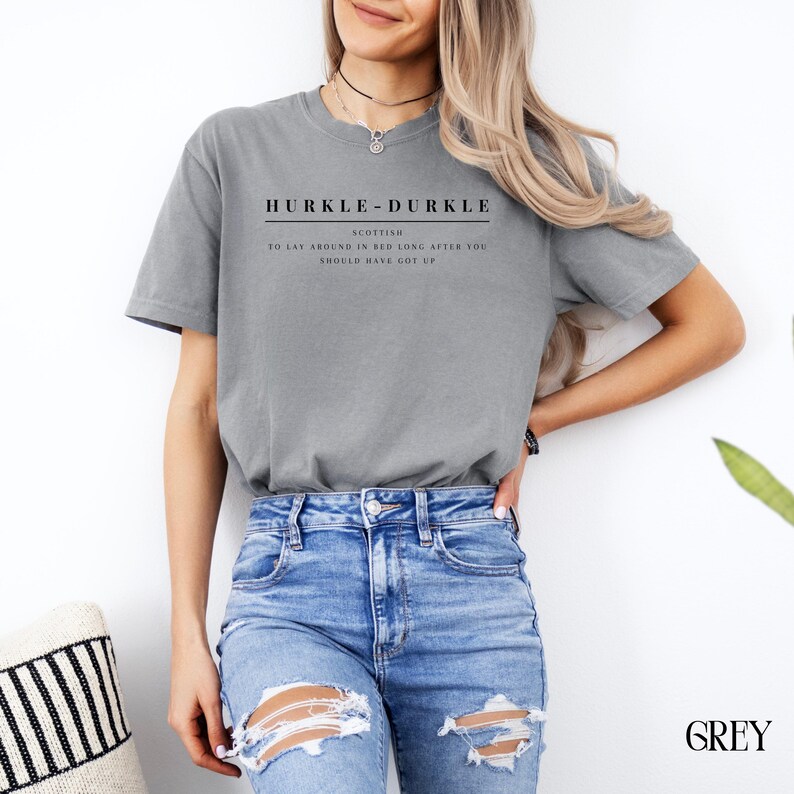 May include: A gray t-shirt with the text "HURKLE-DURKLE" in black, and the words "SCOTTISH", "TO LAY AROUND IN BED LONG AFTER YOU SHOULD HAVE GOT UP" in smaller black text.