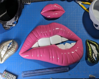 May include: Three pairs of lips, one pink, one silver, and one green, are displayed on a blue surface. The pink lips are large and have white teeth. The silver lips are smaller and have a pearlescent finish. The green lips are also smaller and have a metallic finish.