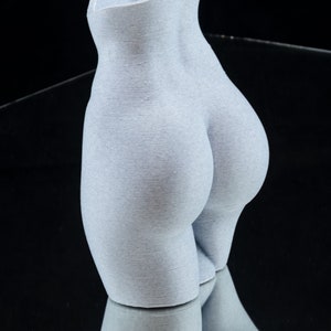 May include: A white ceramic sculpture of a human torso, with a smooth, textured surface. The sculpture is shaped like a human body, with a focus on the hips and buttocks.