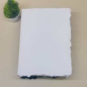 May include: A white, textured paper notebook with a deckled edge. The notebook is lying flat on a light brown surface.