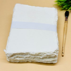 May include: A stack of white handmade paper with deckled edges, bound together with a white ribbon. The paper is arranged on a light brown surface next to a wooden paintbrush.