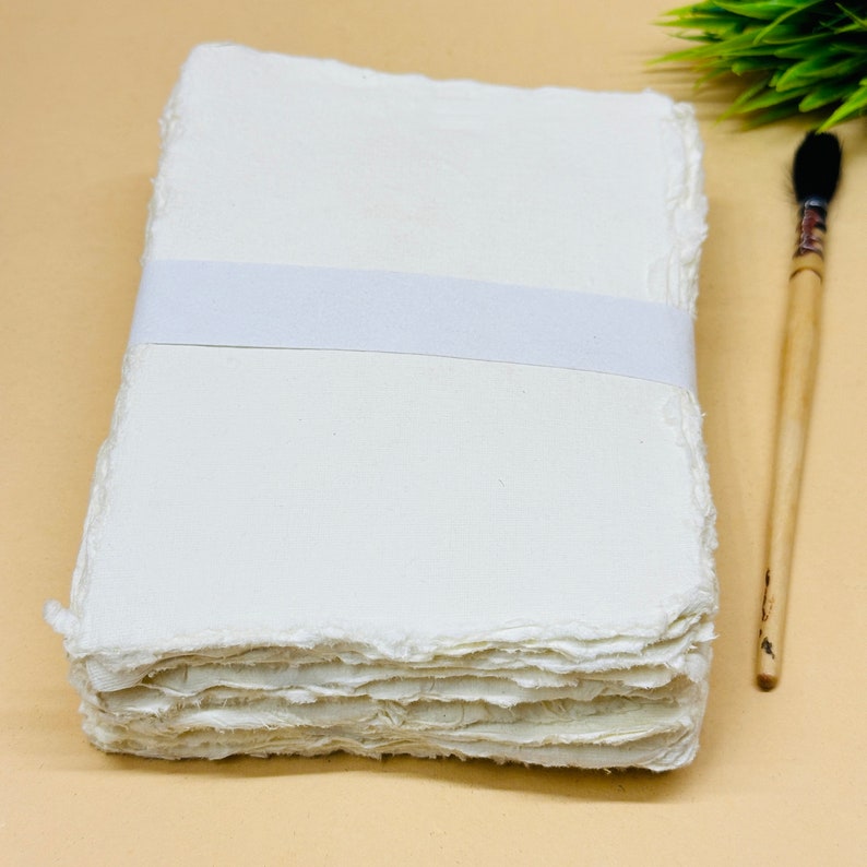 May include: A stack of white handmade paper with deckled edges, bound together with a white ribbon. The paper is arranged on a light brown surface next to a wooden paintbrush.