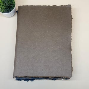 May include: A stack of handmade paper with a brown top sheet and blue bottom sheets. The paper has a rough, textured surface and uneven edges.