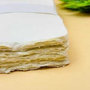 May include: A stack of white handmade paper sheets with a rough, textured edge. The paper is slightly off-white and has a natural, organic look.
