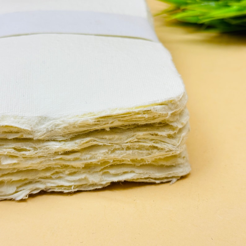 May include: A stack of white handmade paper sheets with a rough, textured edge. The paper is slightly off-white and has a natural, organic look.