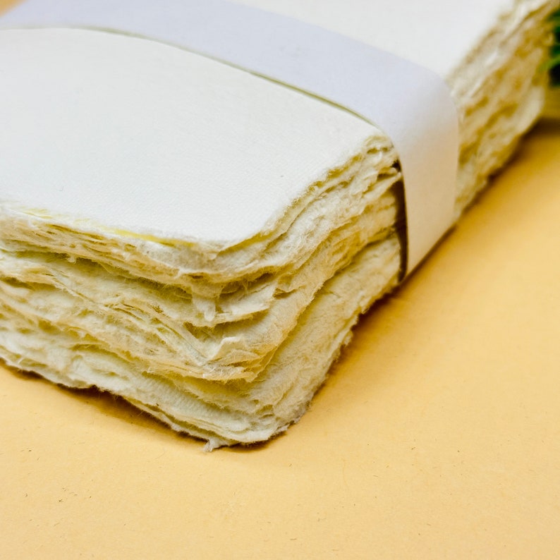 May include: A stack of handmade paper sheets, bound together with a white ribbon. The paper has a rough, textured surface and is a light cream colour.