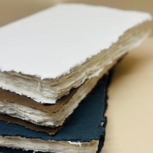 May include: A stack of three handmade paper journals with different colored covers. The top journal has a white cover, the middle journal has a brown cover, and the bottom journal has a dark blue cover.