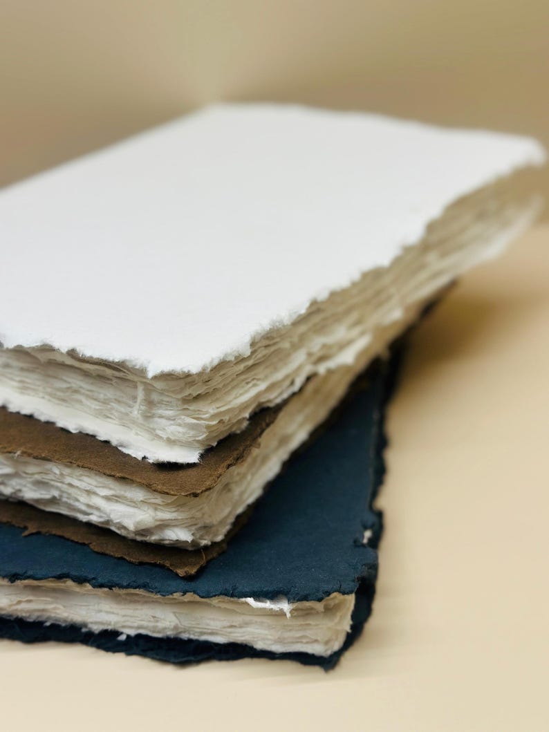 May include: A stack of three handmade paper journals with different colored covers. The top journal has a white cover, the middle journal has a brown cover, and the bottom journal has a dark blue cover.