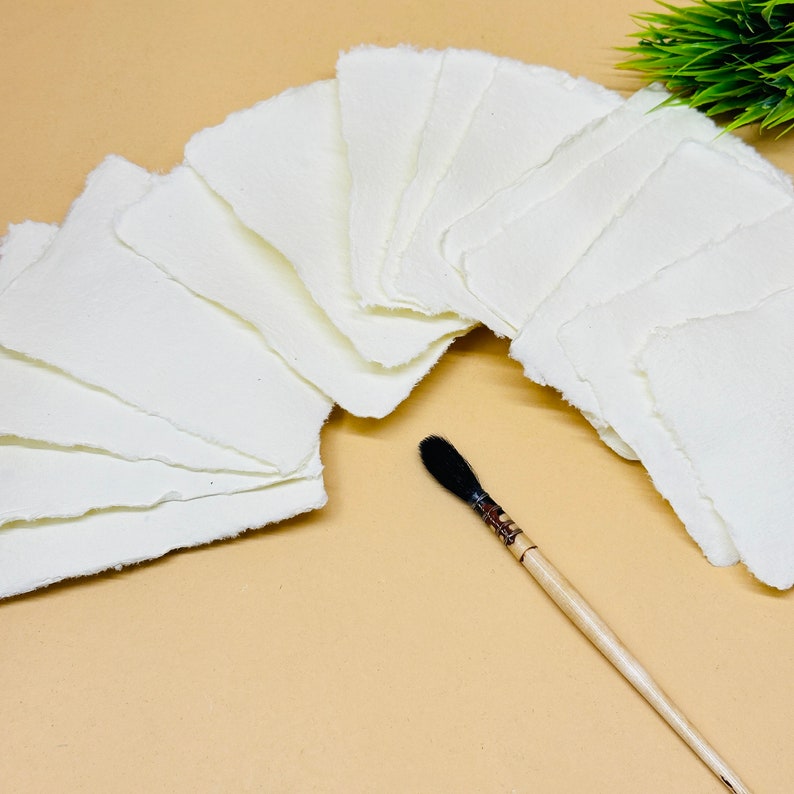 May include: A stack of 15 white handmade paper sheets with deckled edges, arranged in a semi-circle on a beige surface. A black and brown paintbrush is lying on the surface below the paper.