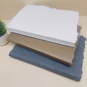 May include: Three notebooks stacked on top of each other. The top notebook is white, the middle notebook is brown, and the bottom notebook is dark blue. All three notebooks have rough, uneven edges.