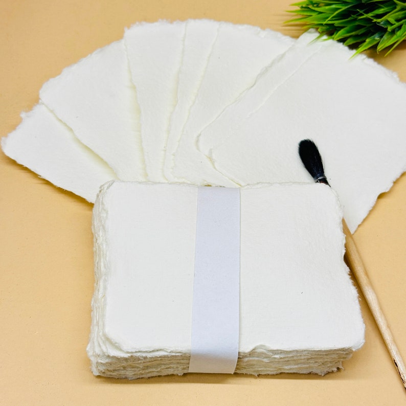 May include: A stack of white handmade paper sheets with deckled edges, tied together with a white ribbon. The paper is arranged in a fan shape, with a black paintbrush and a wooden handle lying on top.