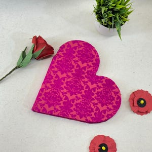 Puede incluir: Una caja de corazón rosa con un patrón floral. La caja está rodeada de flores de papel rojo.