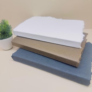May include: Three notebooks stacked on top of each other. The top notebook is white, the middle notebook is brown, and the bottom notebook is dark blue.