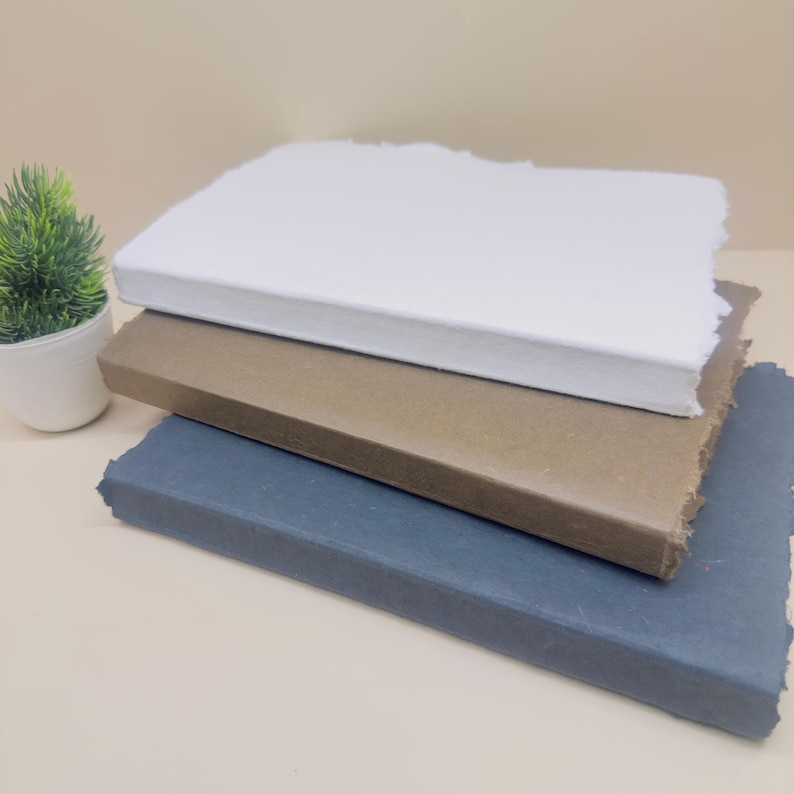 May include: Three notebooks stacked on top of each other. The top notebook is white, the middle notebook is brown, and the bottom notebook is dark blue.
