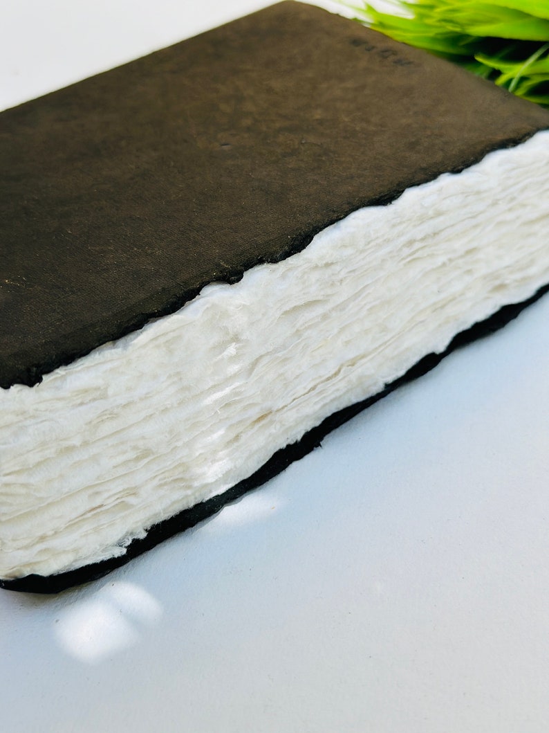 May include: A black leather-bound journal with a stack of blank white pages. The journal is closed and lying on a white surface.