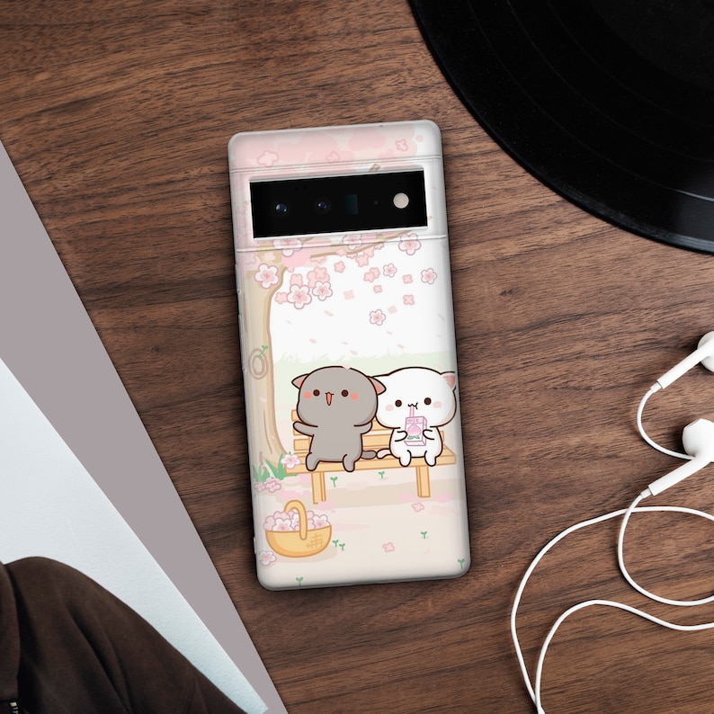 Puede incluir: Una funda de tel&eacute;fono blanca con un dise&ntilde;o de cerezo rosa y blanco. Dos gatos de dibujos animados est&aacute;n sentados en un banco debajo del &aacute;rbol, uno es gris y el otro es blanco. El gato blanco est&aacute; sosteniendo una bebida rosa.