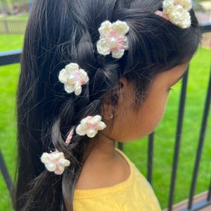 May include: A young girl with long dark hair wearing a yellow shirt and has five white crocheted flower hair clips with pink accents and pearl centers in her hair.