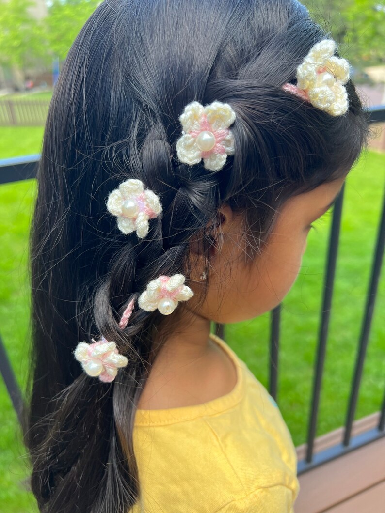 May include: A young girl with long dark hair wearing a yellow shirt and has five white crocheted flower hair clips with pink accents and pearl centers in her hair.