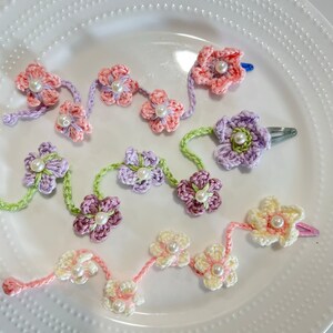 May include: Three crocheted flower hair clips with pearl centers. The clips are in shades of pink, purple, and cream.
