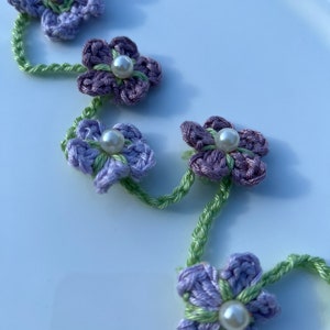 May include: A purple and green crocheted flower hair clip with pearl accents. The clip is made of a chain of flowers connected by green yarn.