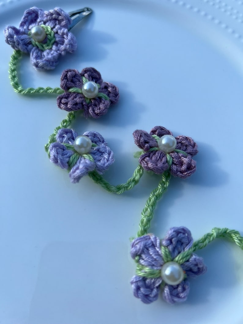 May include: A purple and green crocheted flower hair clip with pearl accents. The clip is made of a chain of flowers connected by green yarn.