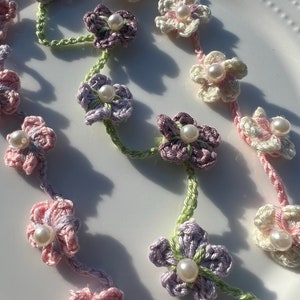 May include: A delicate crocheted flower garland with pastel pink, purple, and white flowers. Each flower has a pearl bead in the center. The garland is strung together with green yarn.