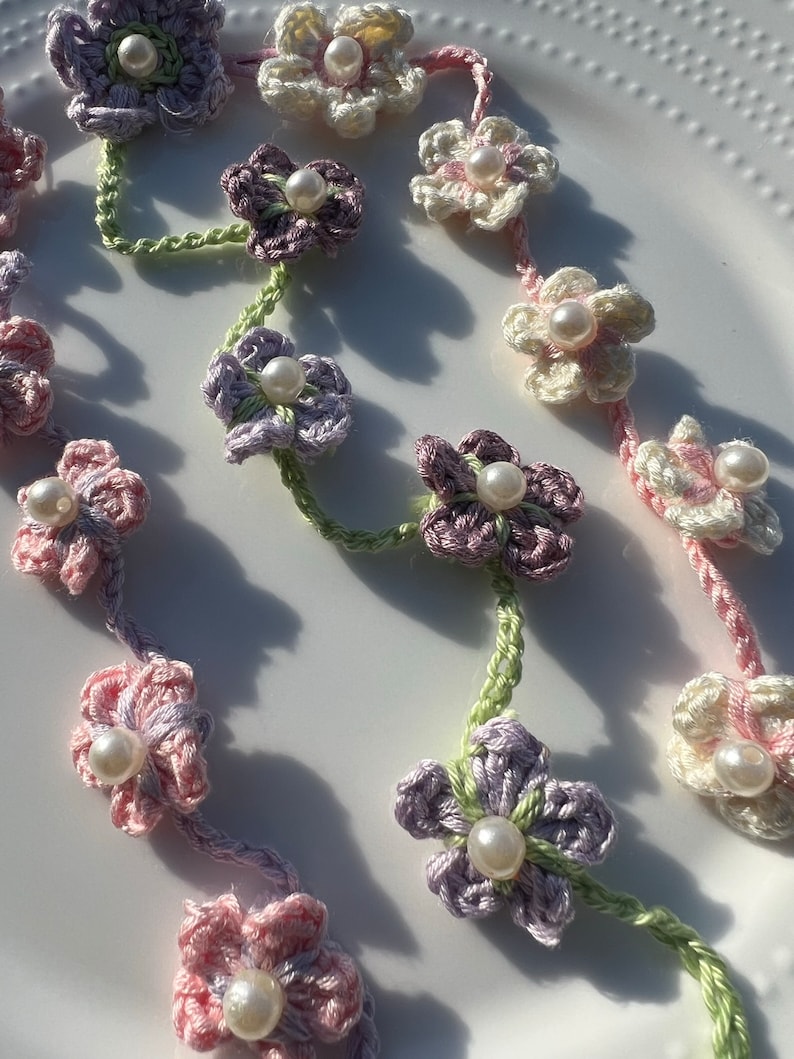May include: A delicate crocheted flower garland with pastel pink, purple, and white flowers. Each flower has a pearl bead in the center. The garland is strung together with green yarn.