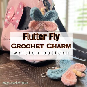Può includere: Un ciondolo a forma di farfalla all'uncinetto rosa e blu con perline di legno. Il ciondolo è attaccato a un portachiavi e pende da una borsa rosa e nera. Il testo "Flutter Fly CROCHET CHARM written pattern" è su uno striscione bianco.