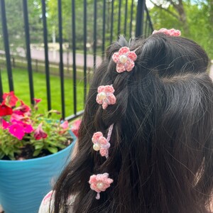 May include: Pink crocheted flower hair clips with a pearl center. The clips are in a girl's hair.