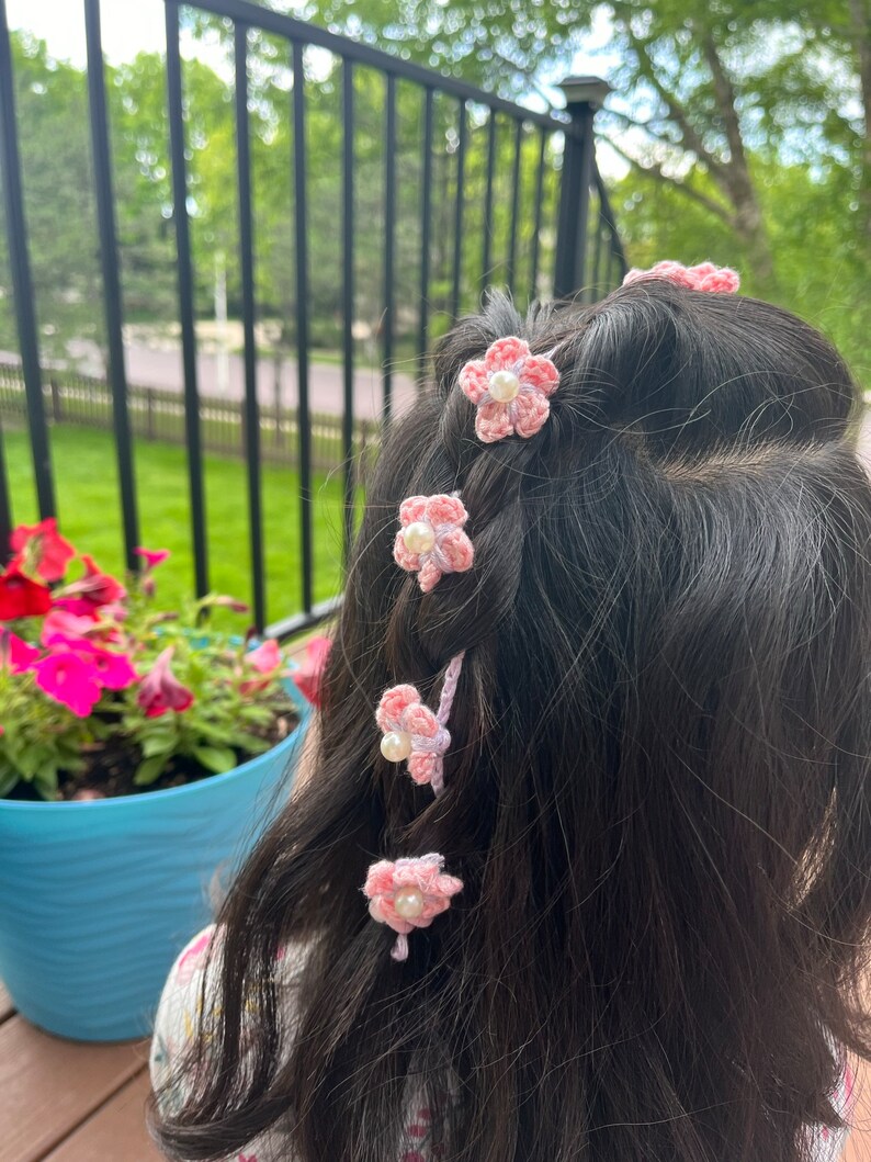 May include: Pink crocheted flower hair clips with a pearl center. The clips are in a girl's hair.