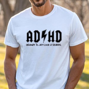 May include: White t-shirt with the text "ADHD" in a black, bold, rock-and-roll style font, with a lightning bolt graphic. Below the text is the phrase "Highway to... Hey look a squirrel" in a smaller font.
