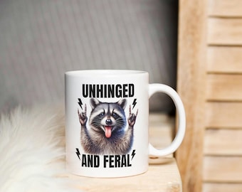 Può includere: Tazza in ceramica bianca con un'illustrazione di procione e la scritta "UNHINGED AND FERAL". Il procione ha la lingua di fuori e fa un gesto rock and roll. La tazza è su una superficie di legno.