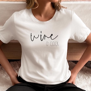 May include: White crew neck t-shirt with the words "wine o' clock" in black script. The shirt is made of a soft material and is suitable for casual wear. The text is centered on the front of the shirt.