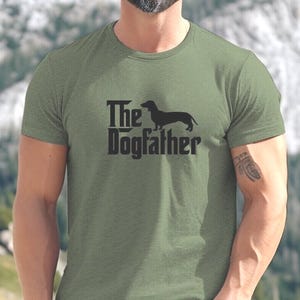May include: An olive green t-shirt featuring the words "The Dogfather" and a dachshund dog silhouette. The t-shirt is made of a soft material and is suitable for casual wear. A great gift for dog lovers.