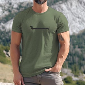 May include: Olive green t-shirt featuring a black dachshund dog graphic. The short-sleeved shirt is made of a soft material. The background shows a mountain landscape.