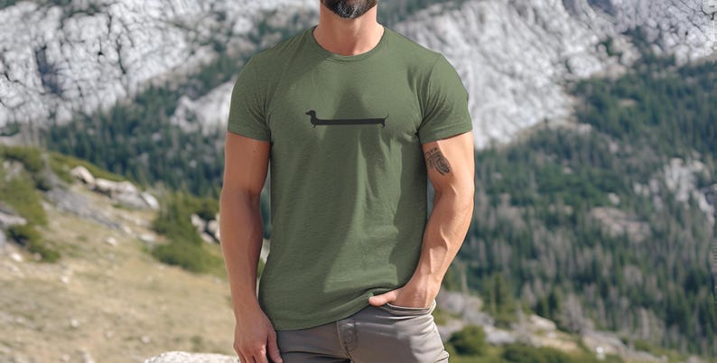 May include: Olive green t-shirt featuring a black dachshund dog graphic. The short-sleeved shirt is made of a soft material. The background shows a mountain landscape.
