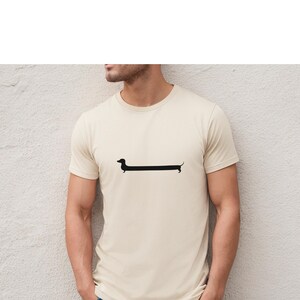 May include: Cream-colored t-shirt featuring a minimalist black dachshund dog graphic. The dog's elongated body is a prominent design element, creating a playful and modern aesthetic. The shirt has short sleeves and a crew neck.