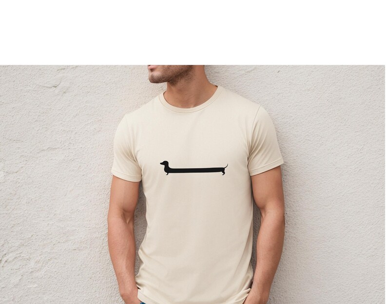 May include: Cream-colored t-shirt featuring a minimalist black dachshund dog graphic. The dog's elongated body is a prominent design element, creating a playful and modern aesthetic. The shirt has short sleeves and a crew neck.