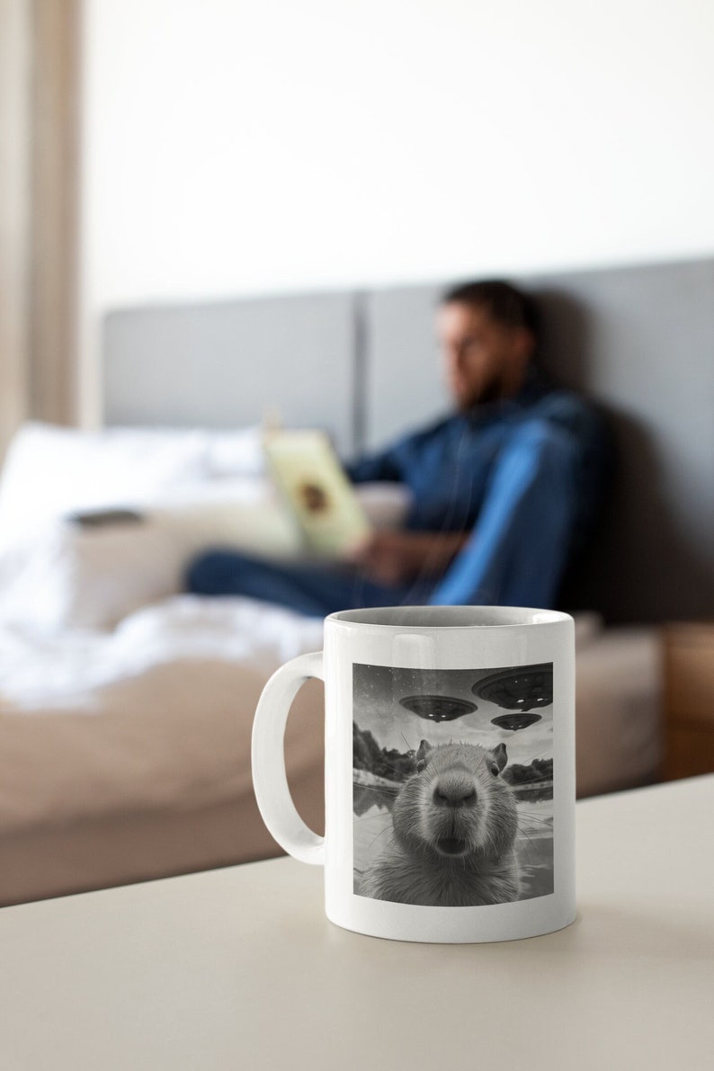 Pu&ograve; includere: Una tazza da caff&egrave; in ceramica bianca con un'immagine in bianco e nero di un capibara che guarda la fotocamera. Il capibara &egrave; in un specchio d'acqua con tre dischi volanti nel cielo sopra.